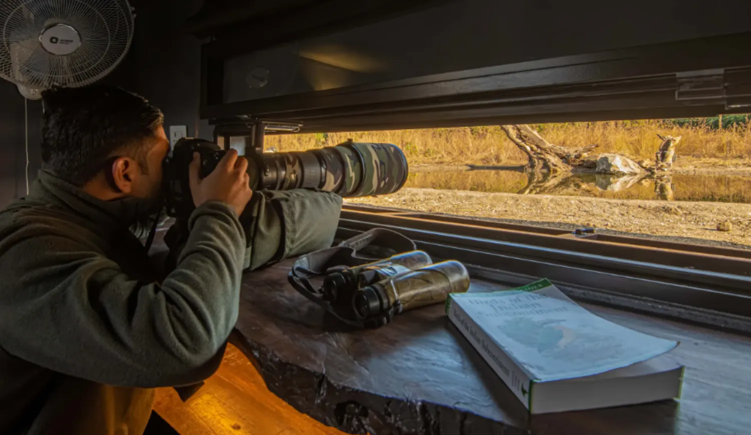 A man using a large telephoto lens camera from inside a hide, looking out over a body of water with a book and binoculars on the ledge.